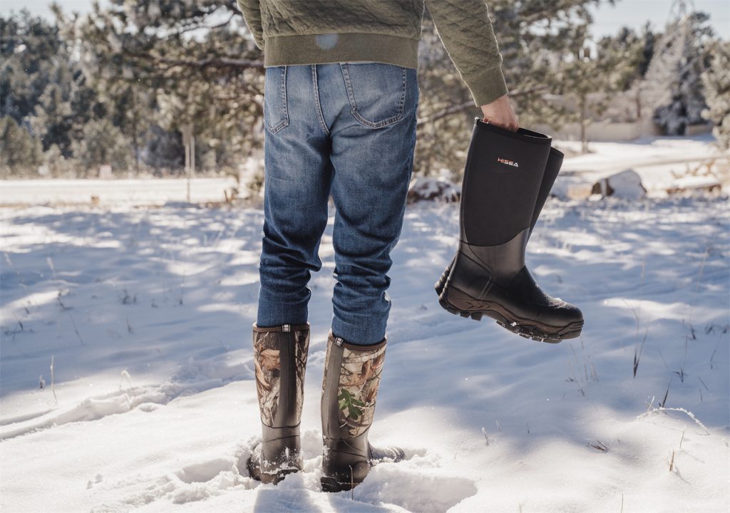 A man is standing in the forest and holding a pair of HISEA snow boots.